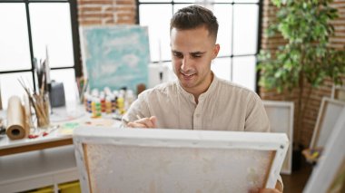 A young hispanic man paints attentively on a canvas, surrounded by art supplies in a bright studio setting. clipart