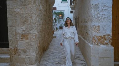 Beyaz elbiseli güzel bir İspanyol kadın polignano 'nun taş döşeli sokaklarında yürüyor ve gülümsüyor bir kısrak, puglia, İtalya.