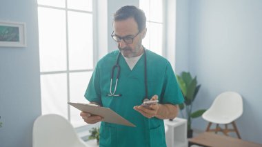 Tıbbi önlüklü olgun İspanyol bir adam bir klinikte pano ve telefonla duruyor, sağlık hizmetleri profesyonelliğini simgeliyor..