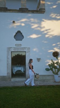 Genç İspanyol bir kadın güneşli bir öğleden sonra Avrupa 'da, Puglia' daki villanın beyaz cephesine karşı bir sepet çanta taşıyan İtalyan masserisine doğru yürür..