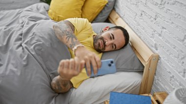 Sakallı bir adam, rahat bir yatak odasında, hafif tuğladan bir arka planı olan bir akıllı telefon kullanarak yatakta dinlenir..