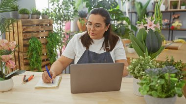 Çiçekçide dizüstü bilgisayarın yanına notlar yazan yetişkin bir kadın.