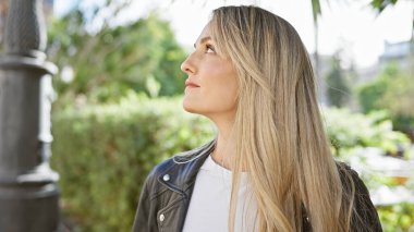 Yeşil bir şehir parkında, düşünceli genç bir kadın, sakin bir ifadeyle yukarı bakıyor..