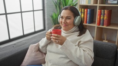 Memnun bir kadın, rahat bir oturma odasında kırmızı kupa ve müzikten zevk alır. Rahatlık ve ev sakinliğinin vücut bulmuş halidir..