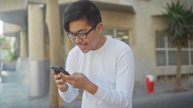 Şehir caddesinde akıllı telefon kullanan Asyalı bir adam gülümsüyor..