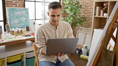 İspanyol adam sanat malzemeleriyle yaratıcı bir stüdyoda dizüstü bilgisayar kullanıyor.