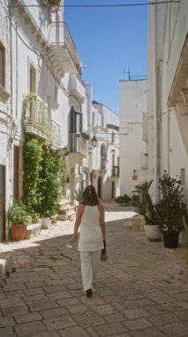 Genç bir İspanyol kadın açık mavi gökyüzünün altında, beyaz binalar ve canlı bitkilerle çevrili Locorotondo, Puglia, İtalya 'nın büyüleyici eski caddelerinde geziniyor..