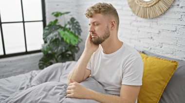 Mavi gözlü ve sakallı dalgın genç bir adam beyaz, modern bir yatak odasında oturuyor.