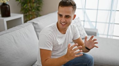 Parlak oturma odasında gülümseyen, beyaz kot pantolon ve saat giyen sıradan İspanyol bir genç.