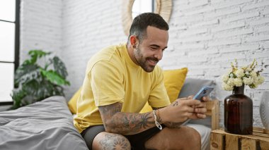 Gülümseyen İspanyol dövmeli genç adam yatak odasında akıllı telefon kullanıyor, rahat ve rahat bir yaşam tarzını şekillendiriyor..