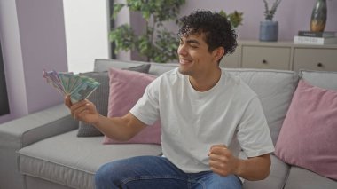 Genç İspanyol bir adam Hindistan rupilerini rahat, modern bir oturma odasında tutarken mutluluk saçar..