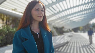 Deniz mavisi ceketli güzel kızıl saçlı bir kadın modern yapıları olan bir şehir parkında dalgın dalgın dolanıyor..