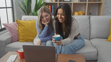 İki kadın kanepede dizüstü bilgisayar ve kahve fincanlarıyla gülümsüyor ve rahatlıyor..