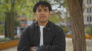 A young handsome hispanic man with curly hair, wearing a black jacket and white shirt, stands with arms crossed in an urban park setting with trees and benches in the background. clipart