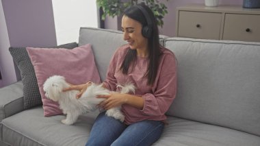 Kulaklıklı gülümseyen bir kadın, rahat bir oturma odasındaki kanepede bir köpeğe evcil hayvan gibi davranır..