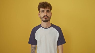 Sakallı ve dövmeli yakışıklı genç bir adam. Parlak sarı bir arkaplanda poz veriyor.