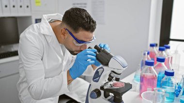 Odaklanmış bir erkek bilim adamı, renkli sıvı örnekleriyle çevrili modern bir laboratuvar ortamında mikroskopla bir numuneyi inceliyor..