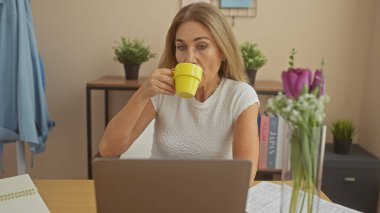 Evde dizüstü bilgisayarla çalışırken kahve içen olgun bir kadın. Etrafı rahat dekor ve bitkilerle çevrili..