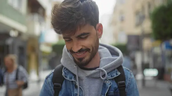 stock image Smiling young hispanic man with beard, wearing denim jacket in urban city street setting.