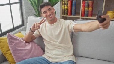 Yakışıklı İspanyol adam oturma odasındaki kanepede akıllı telefonuyla selfie çekiyor, pozitifliğini ve rahatlığını ifade ediyor..