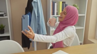 Başörtüsü takan neşeli bir kadın rahat, modern evinde akıllı telefonuyla selfie çekerken kahve fincanından zevk alır..