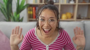 Gülümseyen, gözlüklü, çizgili gömlekli, modern bir oturma odasında oturan İspanyol bir kadın.