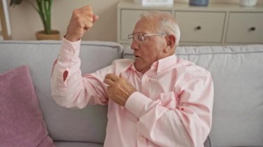 Gri saçlı yaşlı bir adam oturma odasında bir kanepede oturuyor, pembe bir gömlek giyiyor, elini kaldırarak el kol hareketi yapıyor, bir ev, ev, rahat, olgun ve rahat bir atmosfer çiziyor..
