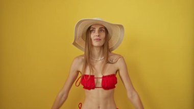 Bikini giyen, şapkasını kaldırıp gülümseyen, izole edilmiş sarı arka planda şaşkın ve mutlu beyaz kadın, neşeyle el kol hareketi yapıyor.
