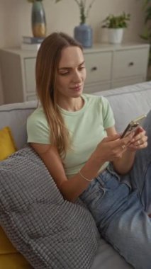 Genç sarışın bir kadın, oturma odasında günlük giysiler giymiş, gri bir koltuğa oturmuş, akıllı telefonuyla nötr bir ifadeyle oturuyor..