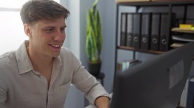 Modern ofiste bilgisayarla çalışan genç adam, gülümseyen ve odaklanmış, organize dosyalar ve bitkilerle çevrili.