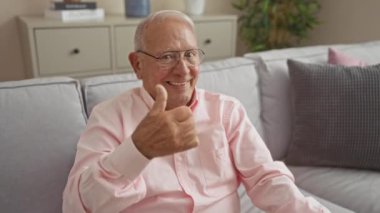 Yaşlı beyaz bir adam, parlak bir oturma odasında, pembe bir gömlek giymiş, rahat gri bir koltukta otururken neşeyle gülümsüyor..