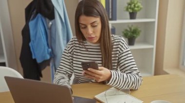Modern bir ofis ortamında akıllı telefon ve laptop kullanan genç İspanyol kadın, uzaktan çalışma ya da dijital iletişimin göstergesi..