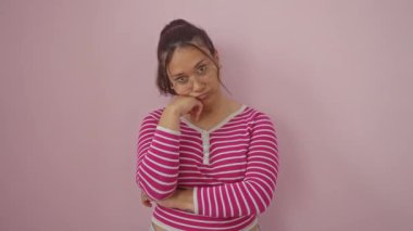 Yorgun görünümlü İspanyol bir kadın, çizgili bir tişört giyiyor, sıkılmış ve depresif duruyor, kollarını kavuşturmuş, sıkıcı düşüncelerde kaybolmuş. Pembe bir duvarın arkasında izole bir şekilde..