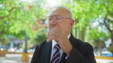 Takım elbiseli ve kravatlı yaşlı bir adam güneşli bir günde canlı bir şehir parkında dikilirken gözlüklerini ayarlıyor..