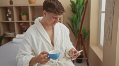 Kaplıcada bir fincan kahvenin tadını çıkaran genç bir adam telefonunu kontrol ederken, arka planda bitki ve rafların bulunduğu sakin, kapalı, sağlıklı bir ortam ile çevrili.
