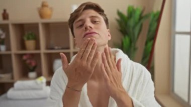 Kaplıca odasında beyaz bir bornoz giyen genç bir adam, bitki ve raflarla dolu bir sağlık merkezinde yüzüne nazikçe dokunuyor..