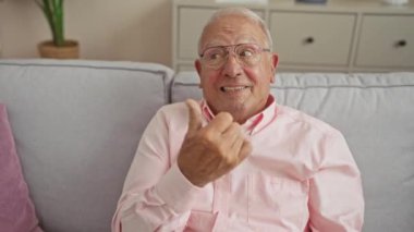 Pembe gömlek giyen, gri saçlı ve gözlüklü neşeli yaşlı bir adam, rahat bir oturma odasında gri bir koltukta oturur, gülümseyerek ve işaret ederek evde oturur..