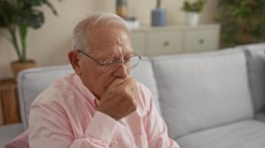 Gri saçlı, gözlüklü ve pembe gömlekli yaşlı bir adam oturma odasında oturuyor, düşünceli görünüyor ve yüzünü elinde tutuyor..