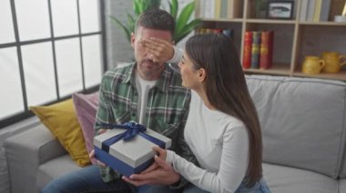 İspanyol kadın, hoş bir oturma odasında hediyesiyle erkeği şaşırtıyor. Sevgi ve neşeyi birlikte ifade ediyor..