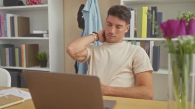 Genç İspanyol bir adam, modern bir oturma odasında dizüstü bilgisayarın başında çalışırken yorgun görünüyor..