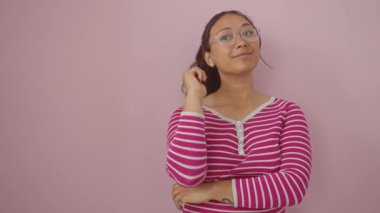 Kendinden emin genç İspanyol kadın, yüzünde ışıl ışıl bir gülümsemeyle gülüyor, pembe izole bir arka plan üzerinde çizgili bir tişörtle dikilirken başka tarafa bakıyor.