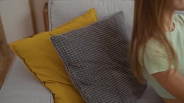 Genç, çekici bir kadın oturma odasında dinleniyor, sarı ve gri yastıklı bir kanepede uzanıyor, rahat bir iç mekanda akıllı telefonu karıştırıyor..