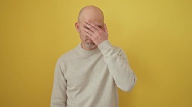 Bald, bearded man in shock, covering his eyes with his hand, fearfully peeking through his fingers. standing against an isolated yellow background, wearing a sweater.