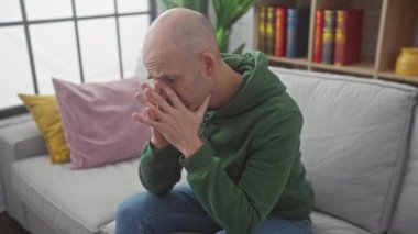 Yeşil kapüşonlu sakallı, stresli kel bir adam. Kapalı bir kanepede oturuyor. Yüzünde ellerle endişeli görünüyor..