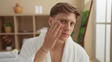 Bir kuaför salonunda cilt bakım ürünleri uygulayan genç bir adam, beyaz bir bornoz giyerek kaplıca ayarlı ve arka planda kapalı bitkiler