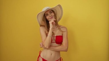 Yazlık şapkalı ve bikinili bir kadın, düşünceli, düşünceli, düşünceli bir yüz ve çene üzerinde eli olan, izole edilmiş sarı bir arka planda, şüpheli bir ifadeyle, fikirlerini sorguluyor.