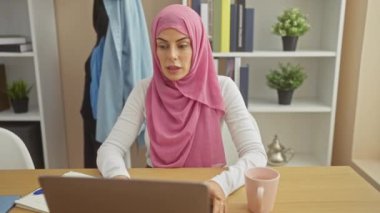 Evde, tesettürlü genç, kendine güvenen İspanyol bir kadın bilgisayarı kullanıyor, elinde dur levhasını cesurca kaldırıyor, kapalı alanda kendini savunmanın ciddi bir ifadesi..