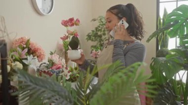 Gülümseyen genç bir İspanyol kadın çiçekçi dükkanında telefonla konuşurken orkideyi inceliyor..