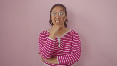 Tırnak yiyen genç İspanyol kadın, pembe arka planda dikiliyor, stresli görünüyor çizgili tişört, korku ve endişe içinde onu rahatsız ediyor.