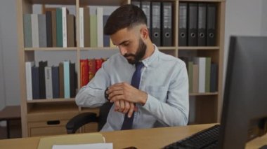 Sakallı, gömlek ve kravat takan genç bir adam, ofis masasında bilgisayarın önünde oturuyor, kol saatine bakıyor, kitap rafları ve evrak işleriyle çevrili.